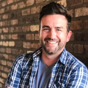 Craig Spilker smiling, wearing a blue plaid shirt in front of a brick wall